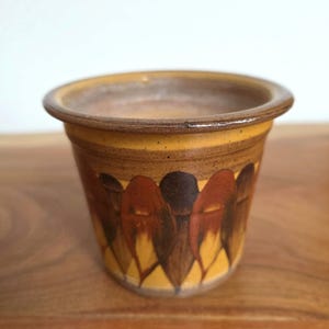 Könnte beinhalten: Ein kleiner Keramiktopf mit einem breiten, abgerundeten Rand. Die Außenseite hat eine warme senfgelbe Grundfarbe, die mit einem sich wiederholenden Muster aus dunkelbraunen und rötlich-orangen tropfenförmigen Elementen verziert ist.