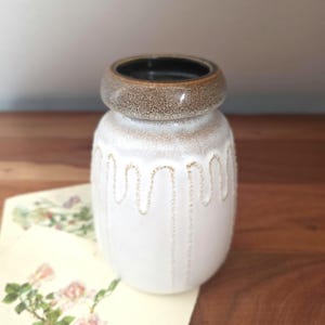 May include: A white ceramic vase with a brown rim and a black interior. The vase has a textured surface with a wavy, dotted design. The vase is sitting on a wooden surface.