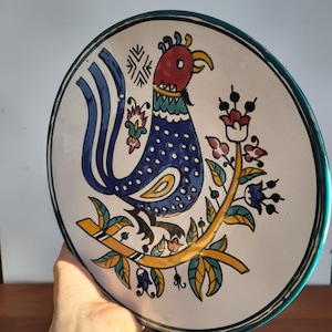 Puede incluir: Un plato de cerámica pintado a mano con un diseño de gallo colorido. El gallo es principalmente azul con detalles rojos, amarillos y verdes. El plato tiene un fondo blanco, un borde verde y motivos florales.