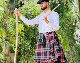 May include: A man wearing a white shirt, black beret, and a red, black, and white plaid kilt. He is holding a wooden staff and standing in a forest setting.