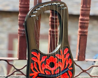 May include: A black lyre with red floral carvings. The instrument has gold strings and is set against a blurred background. The lyre is resting on a concrete surface, showcasing its intricate design and craftsmanship.
