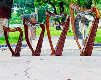 May include: Four brown wooden harps of varying sizes are displayed outdoors. Each harp features intricate carvings and a series of strings. The harps are set against a backdrop of green trees and a concrete surface.