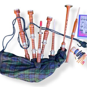 May include: A set of bagpipes with a green and black plaid bag, a chanter, and several drones. The bagpipes are laid out on a white surface with a small booklet titled "The Piper's Guide".