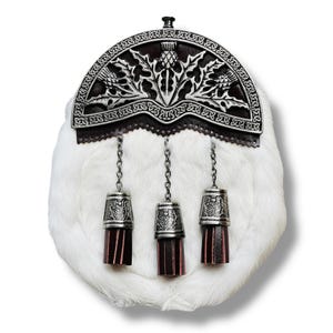 May include: A white fur sporran with a decorative silver metal cantle featuring a thistle design. Three silver tassels with burgundy leather bottoms hang from the sporran, attached by silver chains. The sporran is a traditional Scottish accessory.