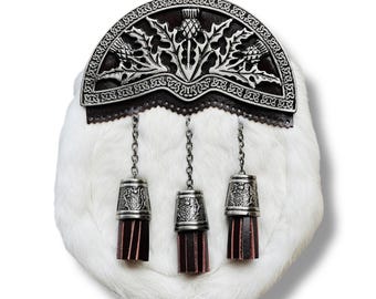 Puede incluir: Una sporran de piel blanca con un cantle decorativo de metal plateado con un diseño de cardo. Tres borlas plateadas con fondos de cuero burdeos cuelgan de la sporran, sujetas por cadenas plateadas. Un accesorio escocés tradicional.