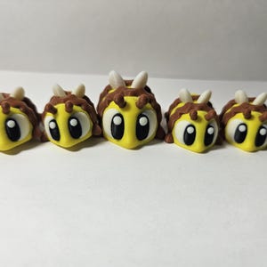 May include: Five yellow and brown bee figurines with large black eyes and white antennae are arranged in a row. The bees have a cartoonish design, with a yellow body and brown stripes. The background is plain white.