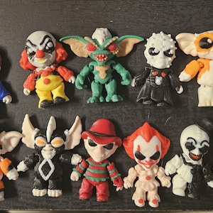 Peut inclure: Une collection de figurines miniatures de personnages de films d'horreur. Les figurines incluent Jason Voorhees, Michael Myers, Pennywise, et d'autres. Chaque figurine est de couleur vive et a un design unique, avec un style de dessin animé.