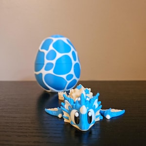 May include: A blue and white dragon figurine with large eyes and a matching egg. The dragon has a spiky crest and fins. The egg is blue with white irregular shapes. The objects are on a dark surface.