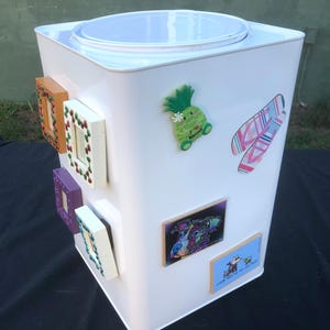 May include: A white rectangular container with a white lid, adorned with various magnets. Magnets include framed pictures, a green pineapple, and colorful surfboard designs. The container is on a black surface.