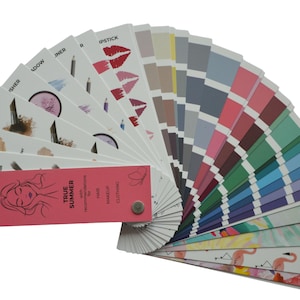 May include: A fan of color swatches for makeup and clothing. The swatches include a range of colors, from neutral to vibrant, and feature illustrations of makeup products. A pink card with text and a woman's face is attached.