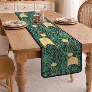 May include: A green and gold leaf pattern table runner with a black border. The runner is placed on a wooden table, with plates and a small wooden tray with salt and pepper shakers. The runner's design features large gold leaves.