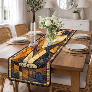 Puede incluir: Un camino de mesa rectangular con un diseño de gato de vidrieras. El camino presenta un mosaico de formas geométricas en tonos naranja, amarillo, marrón y azul, con un borde negro. Las patas del gato son visibles al final del camino.