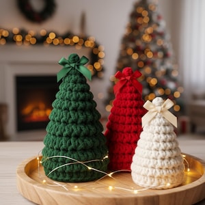 Pode incluir: Três árvores de Natal de crochê em verde, vermelho e creme, cada uma com um laço a condizer. As árvores estão dispostas numa bandeja de madeira com luzes de fio, criando uma exibição festiva.
