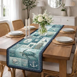 Op de afbeelding: Een rechthoekige tafelloper met een nautisch thema. De loper heeft een patchwork-ontwerp met afbeeldingen van schelpen, zeesterren, ankers en een zeeschildpad in blauw- en wittinten. Het woord "SEA" is gedrukt.