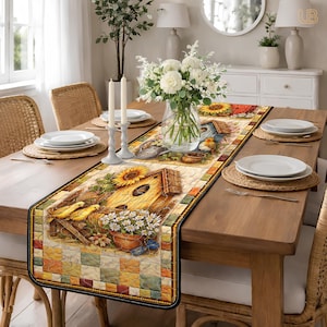 May include: A decorative table runner featuring a birdhouse, sunflowers, and birds. The runner has a quilted design with a black border. The color palette includes yellow, orange, green, and blue. The runner is placed on a wooden table.