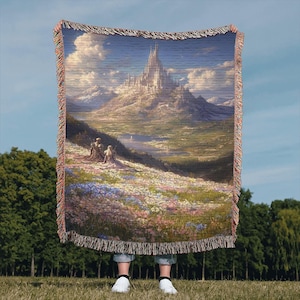 May include: Woven throw blanket depicting a landscape painting of a castle on a mountain, with figures in a field of flowers. The blanket has a fringed edge and is held up against a blue sky.