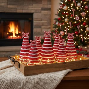 May include: A wooden tray holds seven red and white striped crochet Christmas trees, each topped with a red and white striped bow. The trees are illuminated by small white lights, creating a festive holiday display. A Christmas tree with ornaments is in the background.
