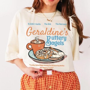 May include: An off-white t-shirt featuring a graphic of a coffee mug and a bagel on a plate with the text Geraldine's Buttery Bagels, R.U.M.P. Castle, The Orb, The Burrow, and Freshly-Baked Bagel Bounty for All Bouncing Bellies and Slippery Treats.