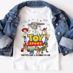May include: White t-shirt featuring the Toy Story characters Buzz Lightyear, Woody, Jessie, and others. The shirt has the words "TOY STORY" in yellow and red, with the phrase "YOU'VE GOT A FRIEND IN ME" below. A blue denim jacket is draped over the shirt.
