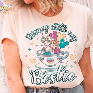 Puede incluir: Camiseta color crema con un gr&aacute;fico de Elsa y Anna de Frozen de Disney en una taza de t&eacute;, con el texto "Disney with my Bestie". El dise&ntilde;o incluye globos coloridos y elementos decorativos.