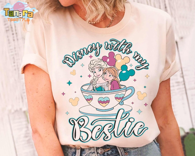 Puede incluir: Camiseta color crema con un gr&aacute;fico de Elsa y Anna de Frozen de Disney en una taza de t&eacute;, con el texto "Disney with my Bestie". El dise&ntilde;o incluye globos coloridos y elementos decorativos.