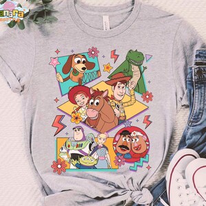Peut inclure: T-shirt gris clair avec un motif color&eacute; des personnages de Toy Story. Le design pr&eacute;sente Woody, Buzz l'&Eacute;clair et d'autres personnages dans un style r&eacute;tro. Le t-shirt est associ&eacute; &agrave; un jean bleu et des baskets blanches.