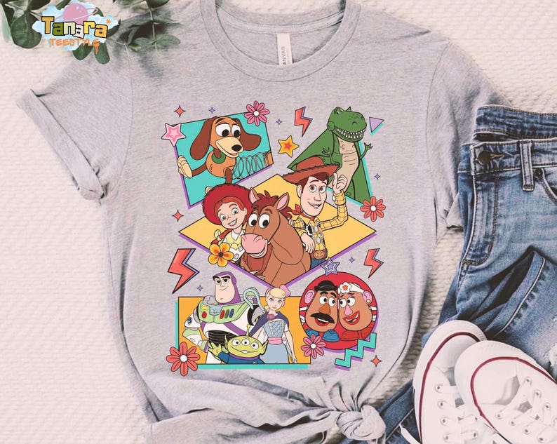 Peut inclure: T-shirt gris clair avec un motif color&eacute; des personnages de Toy Story. Le design pr&eacute;sente Woody, Buzz l'&Eacute;clair et d'autres personnages dans un style r&eacute;tro. Le t-shirt est associ&eacute; &agrave; un jean bleu et des baskets blanches.