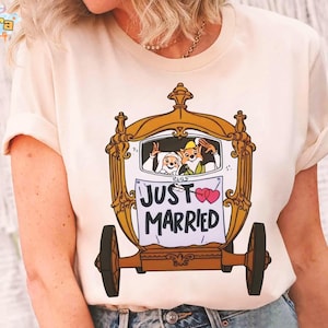 May include: Cream-colored t-shirt featuring a cartoon carriage with the words "JUST MARRIED" and two pink hearts. The carriage has a gold frame and brown wheels. Inside the carriage, cartoon characters wave. The shirt has short sleeves.