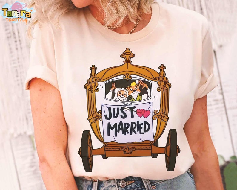 May include: Cream-colored t-shirt featuring a cartoon carriage with the words "JUST MARRIED" and two pink hearts. The carriage has a gold frame and brown wheels. Inside the carriage, cartoon characters wave. The shirt has short sleeves.