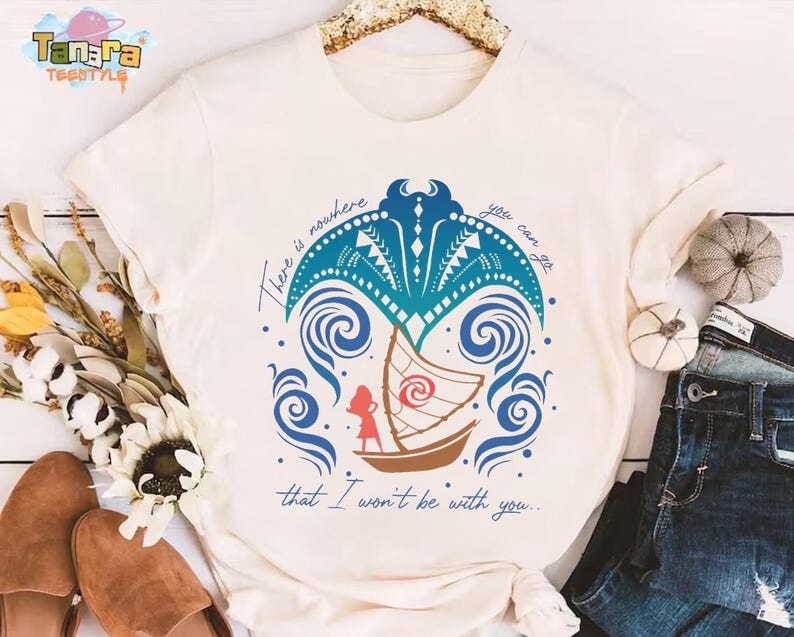 May include: A cream-colored short-sleeve t-shirt featuring a graphic of a brown sailboat, a red silhouette of a person, and a blue manta ray design. The text reads, There is nowhere you can go that I won't be with you, in a blue cursive script.