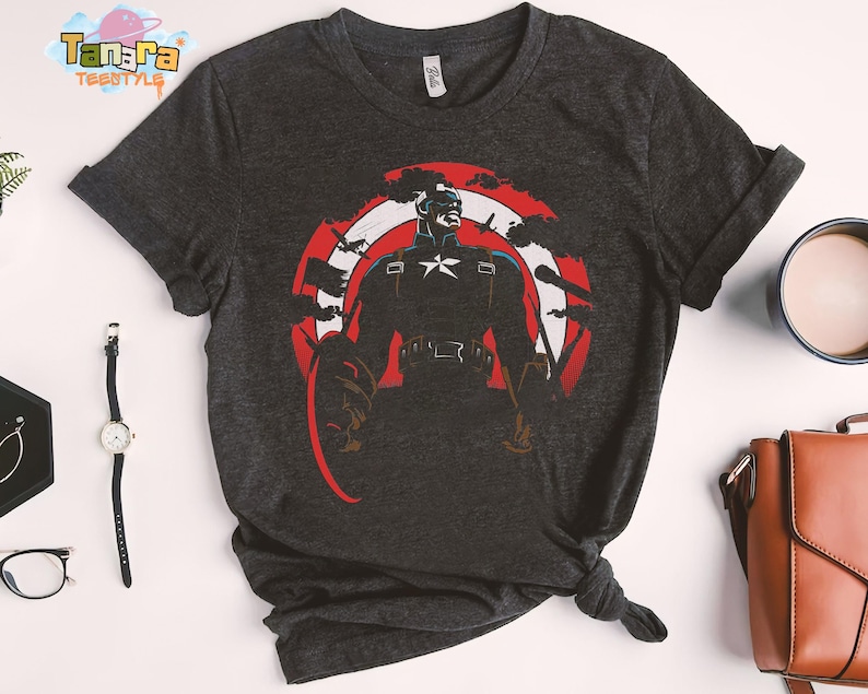 K&ouml;nnte beinhalten: Dunkelgraues T-Shirt mit einem Captain America-Motiv. Das Design zeigt Captain America in einer heldenhaften Pose, vor einem rot-wei&szlig;-blauen Schild. Das T-Shirt hat kurze &Auml;rmel und einen Rundhalsausschnitt.
