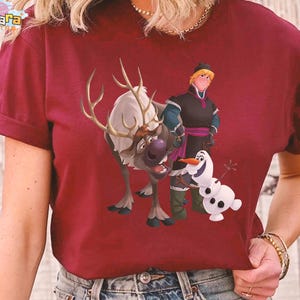 Peut inclure: T-shirt bordeaux avec un graphisme color&eacute; des personnages du film d'animation "La Reine des Neiges". Le motif comprend un renne, un bonhomme de neige et deux personnages humains. Le t-shirt a des manches courtes et un col rond.