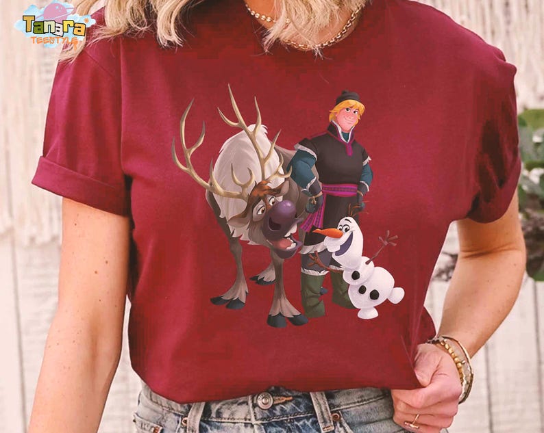 Peut inclure: T-shirt bordeaux avec un graphisme color&eacute; des personnages du film d'animation "La Reine des Neiges". Le motif comprend un renne, un bonhomme de neige et deux personnages humains. Le t-shirt a des manches courtes et un col rond.