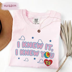 May include: A light pink short-sleeve t-shirt featuring the text I KNEW IT, I KNEW YOU in blue and pink letters. The design includes blue stars, white clouds, a colorful ball with a red star, and a red heart containing the yellow letters TS.