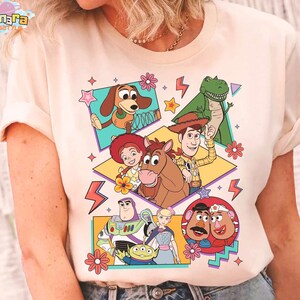 Peut inclure: T-shirt cr&egrave;me avec un graphisme color&eacute; des personnages du film d'animation "Toy Story". Le motif comprend des images de Woody, Buzz l'&Eacute;clair et d'autres personnages, avec des &eacute;toiles, des fleurs et des &eacute;clairs d&eacute;coratifs.