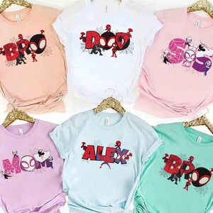 Puede incluir: Camisetas de varios colores con dise&ntilde;os de Spider-Man. Las camisetas presentan las palabras "BRO", "DAD", "MOM", "ALEX" y "BOO" en una fuente estilizada, junto con personajes de Spider-Man y telara&ntilde;as. Las camisetas se muestran en perchas doradas.