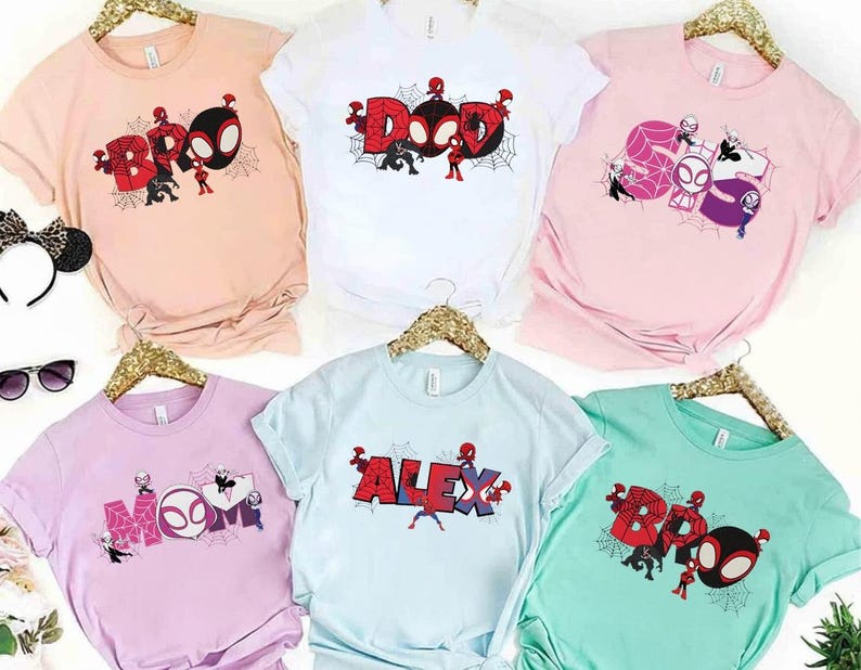 Puede incluir: Camisetas de varios colores con dise&ntilde;os de Spider-Man. Las camisetas presentan las palabras "BRO", "DAD", "MOM", "ALEX" y "BOO" en una fuente estilizada, junto con personajes de Spider-Man y telara&ntilde;as. Las camisetas se muestran en perchas doradas.