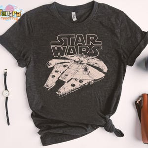 Può includere: Maglietta grigio scuro di Star Wars con il Millennium Falcon bianco e il logo di Star Wars. La maglietta è annodata in basso. Un orologio, occhiali e una tazza di caffè sono accanto alla maglietta.