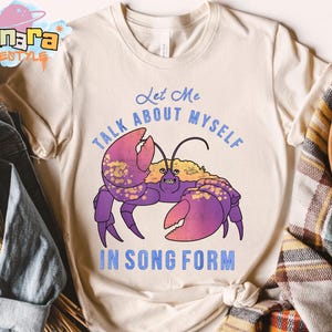 Puede incluir: Camiseta color crema con una ilustraci&oacute;n de cangrejo morado, pinzas rosas y detalles dorados. El texto "Let Me Talk About Myself In Song Form" est&aacute; impreso en azul encima y debajo del cangrejo.