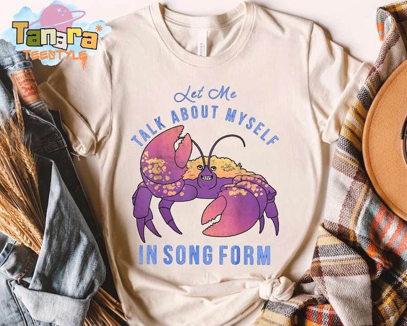 Puede incluir: Camiseta color crema con una ilustraci&oacute;n de cangrejo morado, pinzas rosas y detalles dorados. El texto "Let Me Talk About Myself In Song Form" est&aacute; impreso en azul encima y debajo del cangrejo.