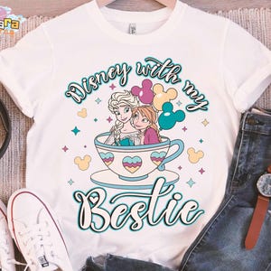 Puede incluir: Camiseta blanca con una ilustraci&oacute;n de dibujos animados de Elsa y Anna de Frozen de Disney, sentadas en una taza de t&eacute; con globos de Mickey Mouse. El texto "Disney with my Bestie" est&aacute; escrito en una fuente caprichosa.