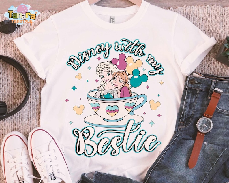 Puede incluir: Camiseta blanca con una ilustraci&oacute;n de dibujos animados de Elsa y Anna de Frozen de Disney, sentadas en una taza de t&eacute; con globos de Mickey Mouse. El texto "Disney with my Bestie" est&aacute; escrito en una fuente caprichosa.