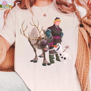 Peut inclure: T-shirt cr&egrave;me avec un graphisme des personnages du film d'animation "La Reine des Neiges". Le motif comprend un renne, un bonhomme de neige et deux personnages humains. La marque "Tanara Tees" est imprim&eacute;e en haut &agrave; gauche.