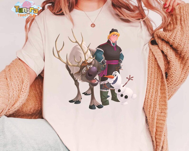 Peut inclure: T-shirt cr&egrave;me avec un graphisme des personnages du film d'animation "La Reine des Neiges". Le motif comprend un renne, un bonhomme de neige et deux personnages humains. La marque "Tanara Tees" est imprim&eacute;e en haut &agrave; gauche.