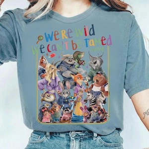May include: A light blue t-shirt featuring a colorful graphic of various cartoon characters. The text "we're wild we can't be tamed" is above the characters. The shirt is made of a soft material.