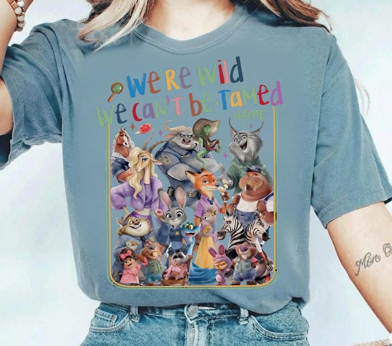 May include: A light blue t-shirt featuring a colorful graphic of various cartoon characters. The text "we're wild we can't be tamed" is above the characters. The shirt is made of a soft material.