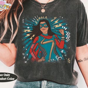 Op de afbeelding: Donkergrijs t-shirt met een afbeelding van een superheld, met de naam "KAMALA" erboven. De superheld draagt een blauw masker, rode cape en een blauw en rood pak. De achtergrond heeft blauwe stralen en bliksem.