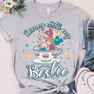 Puede incluir: Camiseta gris claro con un gr&aacute;fico colorido de dos personajes animados en una taza de t&eacute;, con el texto "Disney with my Bestie". El dise&ntilde;o incluye corazones, estrellas e iconos de Mickey Mouse.