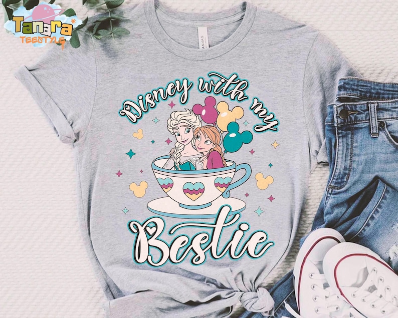 Puede incluir: Camiseta gris claro con un gr&aacute;fico colorido de dos personajes animados en una taza de t&eacute;, con el texto "Disney with my Bestie". El dise&ntilde;o incluye corazones, estrellas e iconos de Mickey Mouse.