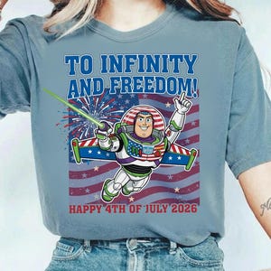 May include: Light blue t-shirt featuring Buzz Lightyear with the text "TO INFINITY AND FREEDOM!" and "HAPPY 4TH OF JULY 2026" over an American flag design. Buzz is holding a green laser and pointing upwards.